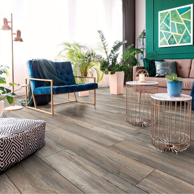 Unser Mittelklasse-Laminat Raumfoto eines Laminats in grauer Holzoptik in einem modernen Wohnzimmer in den Farben Hellbraun, Rosé und Grün. Im Hintergrund sind Tische und ein Stuhl zu erkennen.