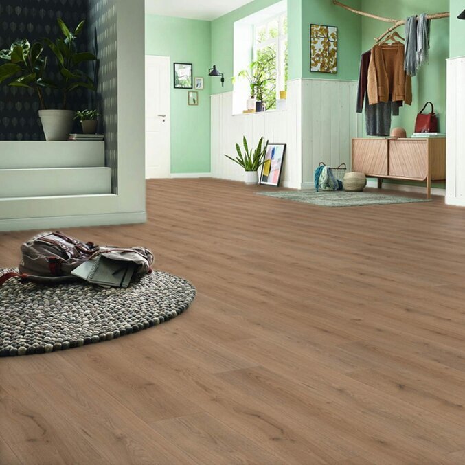 Unser Basic-Laminat Raumfoto eines Laminats in brauner Holzoptik in einem offenen Wohnbereichen in den Farben Braun und Grün. Im Hintergrund ist eine Treppe zu erkennen, im Vordergrund ein grauer Teppich.